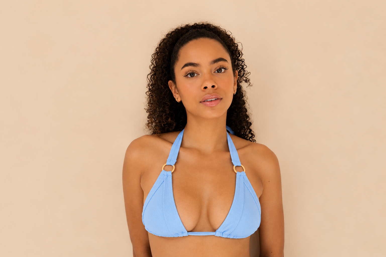 A woman wearing a light blue triangle bikini top with gold-tone ring details on the straps, posed against a solid beige background.